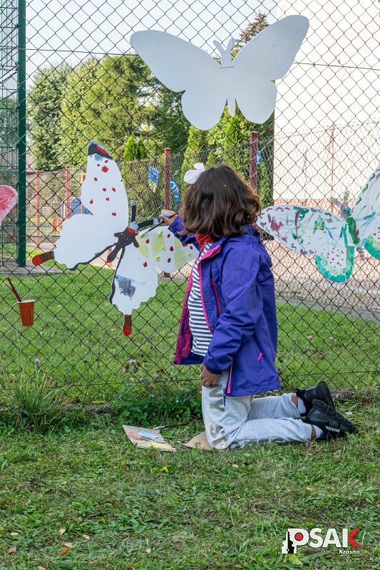 Dziewczynka klęczy i maluje białe motyle zawieszone na ogrodzeniu