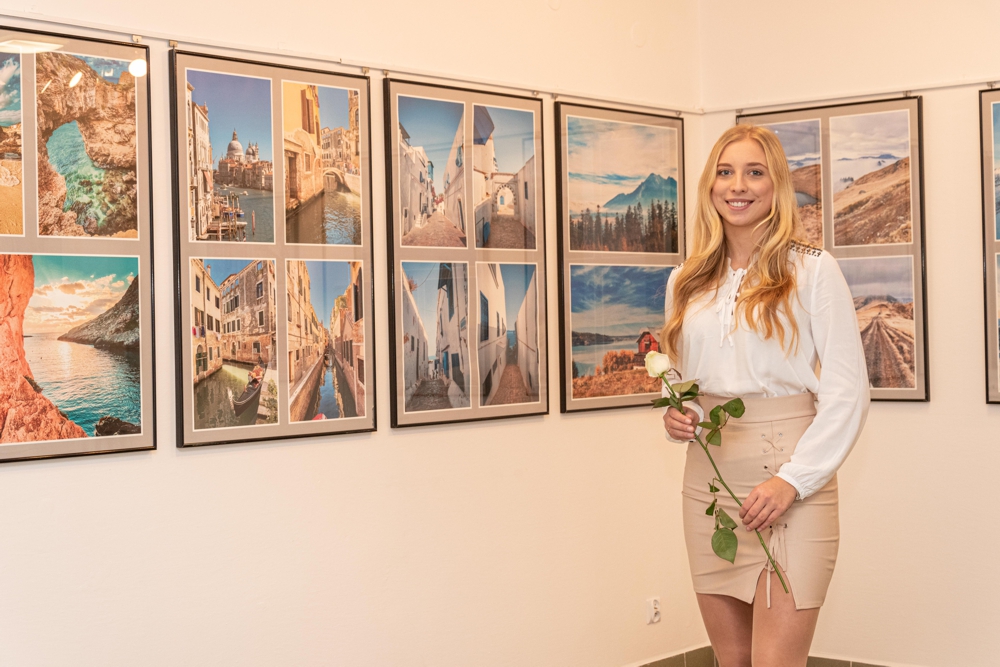  Na zdjęciu autorka wystawy fotograficznej  Klaudia Glazar stojąca na tle swoich zdjęć, które przedstawiają krajobrazy. Autorka w rękach trzyma białą różę.