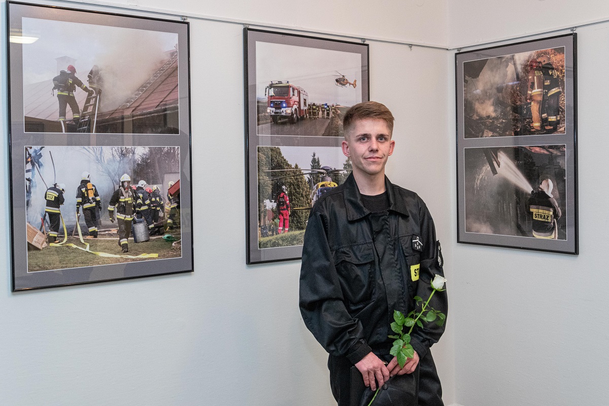 Autor jednej z wystaw fotograficznych stojący na tle swoich zdjęć. W rękach trzyma białą różę.