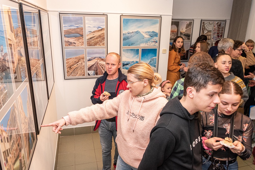 Zdjęcie przedstawia widzów oglądających wystawę fotograficzną Klaudii Glazar. Na ścianach wiszą zdjęcia z krajobrazami. 