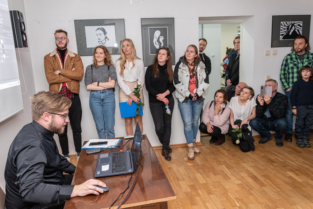 Na zdjęciu autor wystawy Hubert Żak siedzi przy biurku z laptopem i przestawia prezentację. Obok stoją widzowie. Na ścianach wiszą prace autora. 